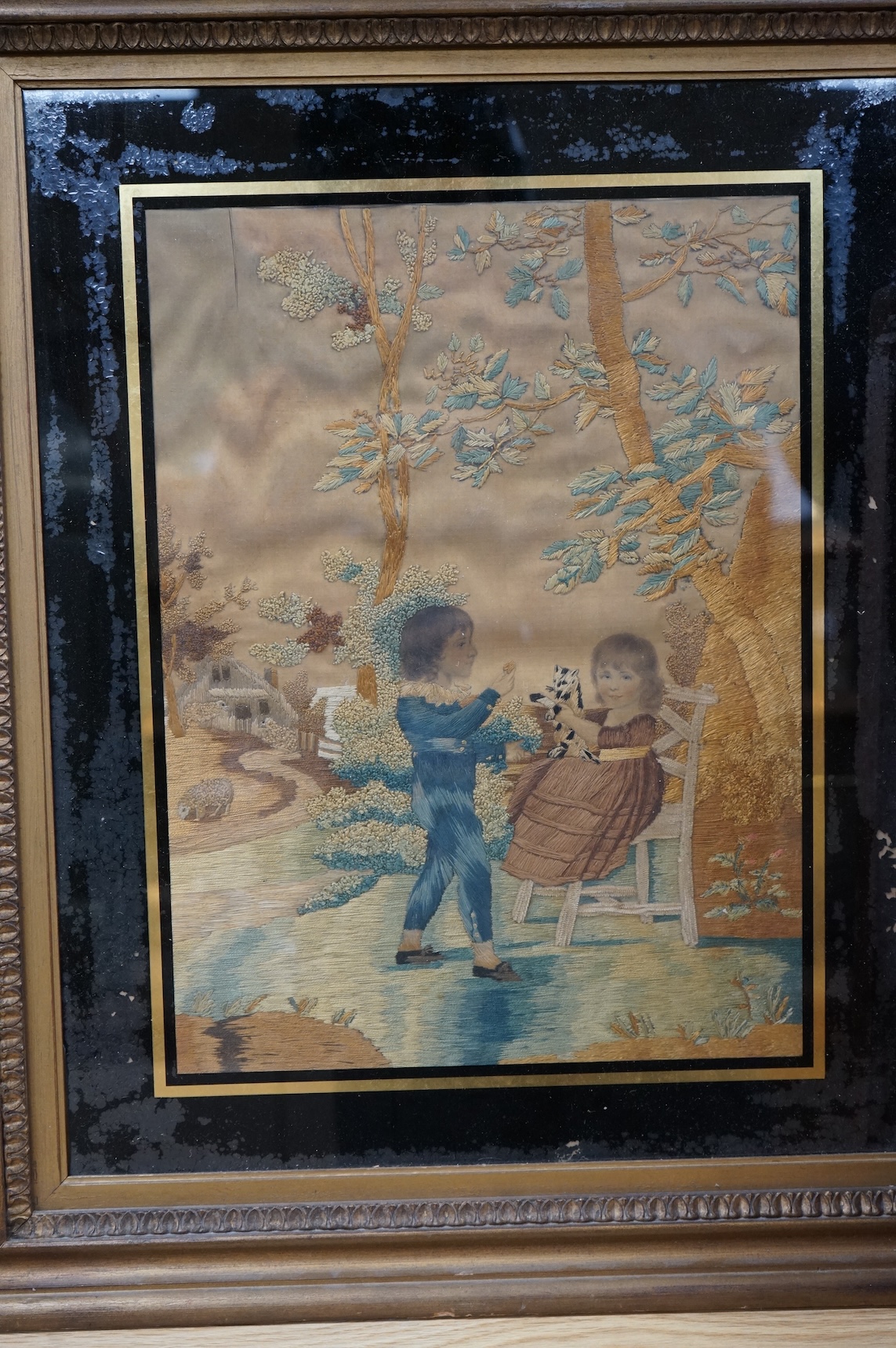 Two early 19th century silk worked embroideries one of two children with a cat, the other of a seated lady reading. Both within Verre Eglomise glass and framed. Largest 32cm x 24cm.
